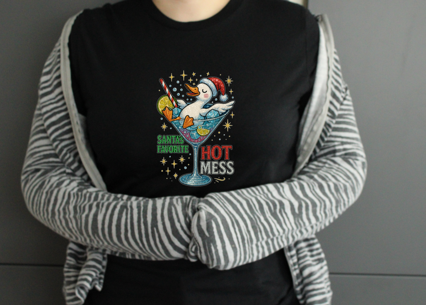 Person wearing a black tshirt with a colourful graphic design duck in cocktail glass saying santas hot mess on a plain background
