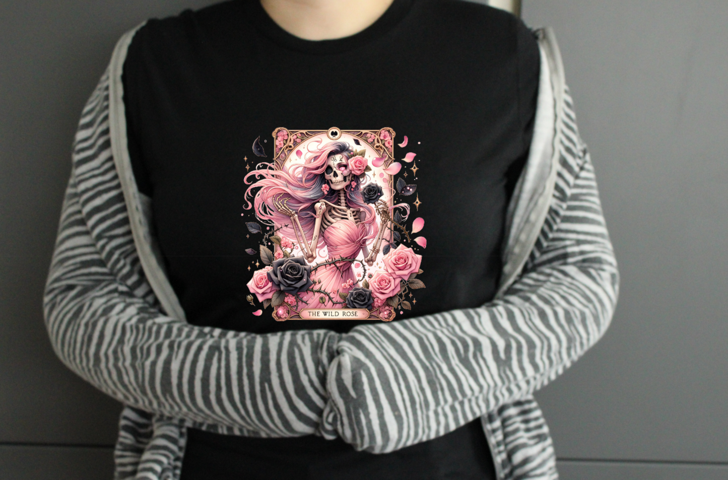 Person wearing a black tshirt with a pink and white wild rose tarot card witchy design, standing against a gray background.