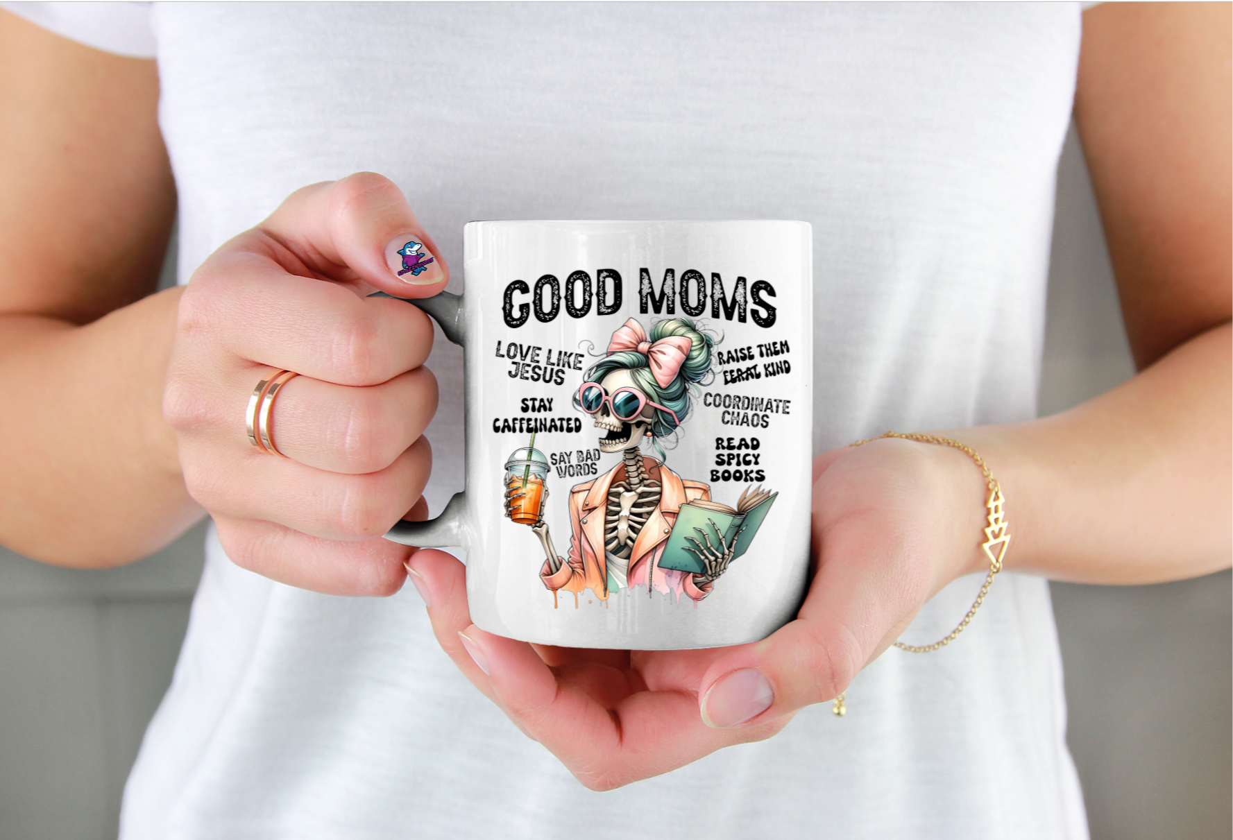 Person holding a mug with a skull design and text Good Moms Say Bad Words, wearing a white shirt.