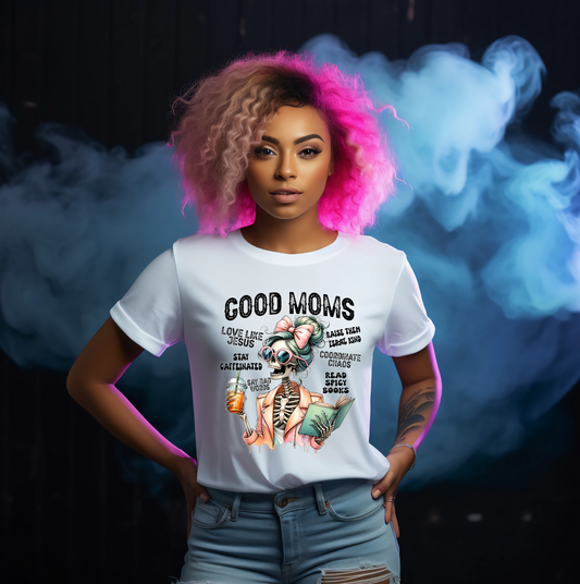 Person wearing a white t-shirt with graphic design and text Saying Good Moms Say Bad Words with gothic skeleton graphic, standing against a dark background with smoke effects.