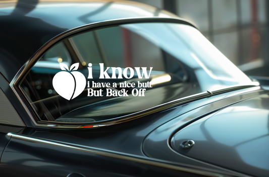 Car window with a humorous decal reading 'i know I have a nice butt But Back Off' on a reflective surface.